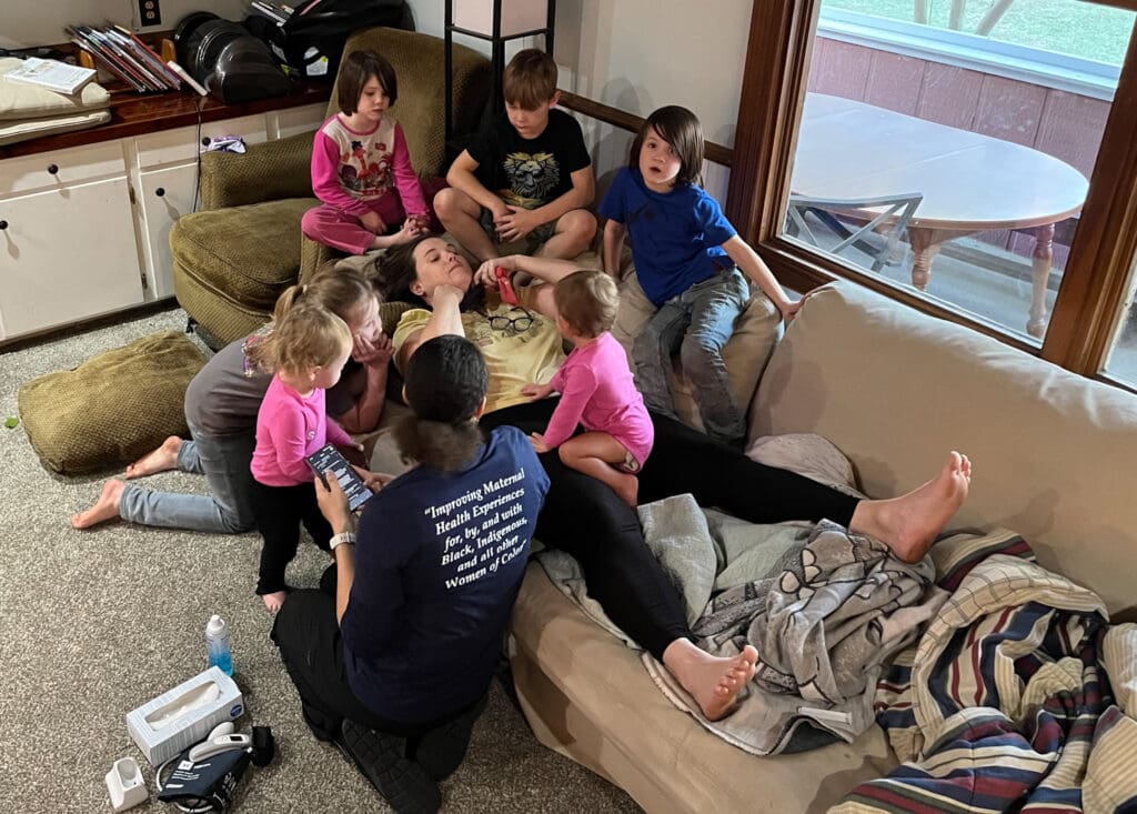 Tifton birth photographer: Mom lays on couch, surrounded by kids, while midwife monitors baby