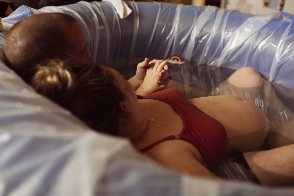 Understanding the cost of birth photoraphy in Tifton: sweet moment, pregnant woman is leaning back against her husband in birthing pool, the image is taken from behind them and focuses on their holding hands