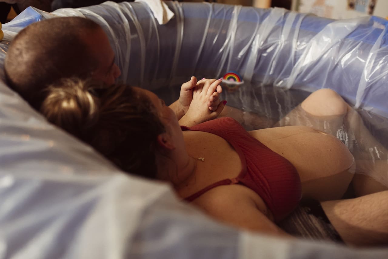 Understanding the cost of birth photoraphy in Tifton: sweet moment, pregnant woman is leaning back against her husband in birthing pool, the image is taken from behind them and focuses on their holding hands