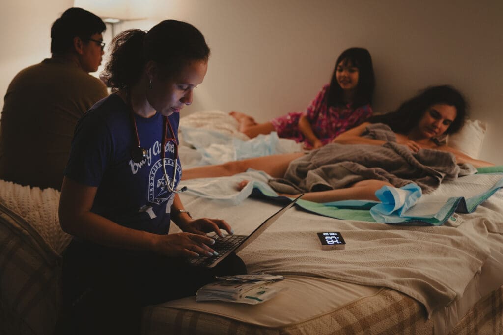Nurse midwife Victoria Gordon working on her laptop after delivering a baby at a home birth while family rests and enjoys newborn in the background, by Tifton birth & newborn photographer