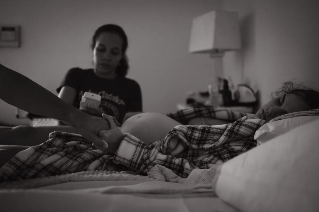 Why more families are choosing home birth: midwife listens to baby's heartbeat on doppler while Mom rests on the bed, holding husband's hand for comfort
